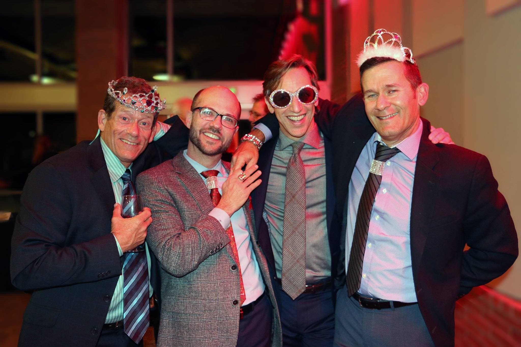 Four guests in suits laughing together at a gala, two wearing tiaras and one in rhinestone sunglasses.