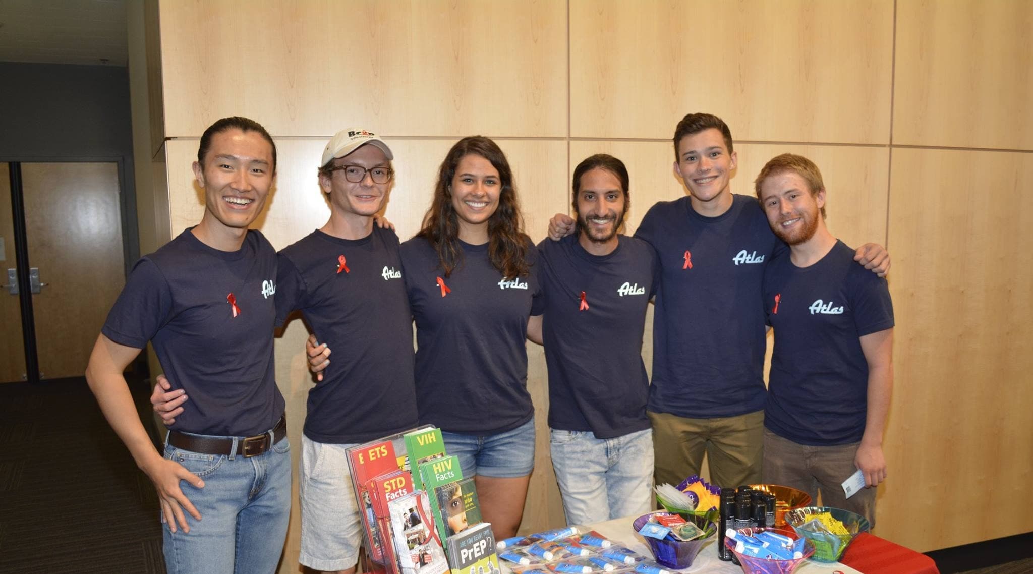 Smiling BCAP outreach volunteers in navy Atlas t-shirts, most wearing red ribbon pins, behind a table of HIV Facts and PrEP brochures.